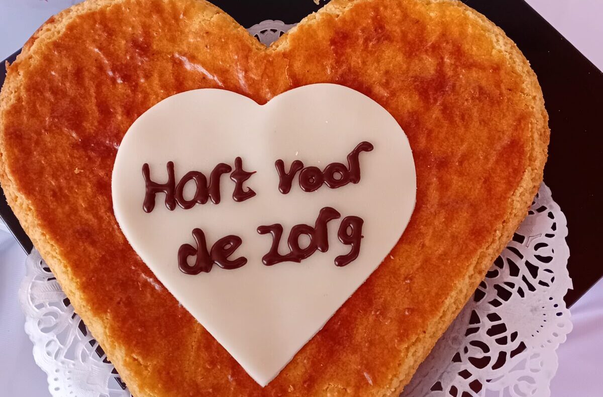 Hartvormige taart met een witte fondantcirkel en de tekst "Hart voor de zorg" erop, op een kanten onderzetter.