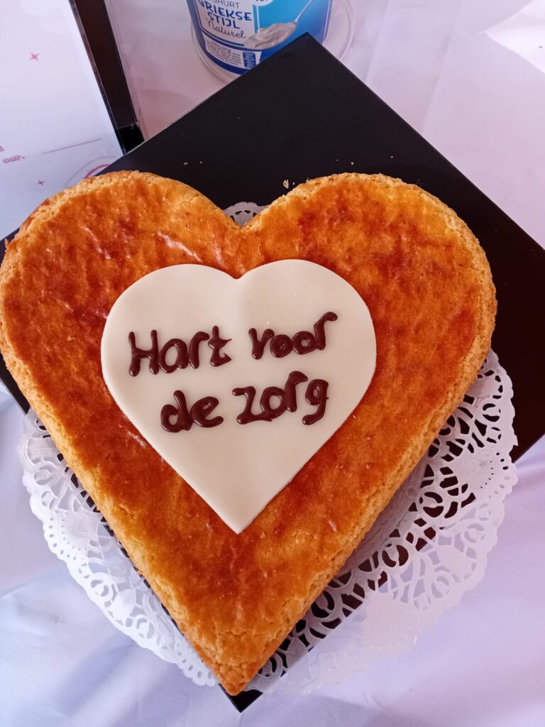 Hartvormige taart met een witte fondantcirkel en de tekst "Hart voor de zorg" erop, op een kanten onderzetter.