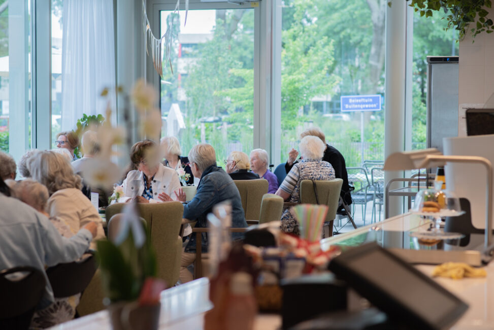 Groep ouderen zit samen in een lichte ruimte met grote ramen, omringd door groen. Ze zijn in gesprek en genieten van de omgeving.
