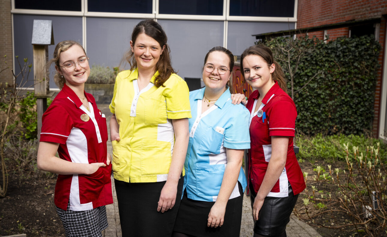 Vier vrouwen in kleurrijke uniformen staan lachend buiten voor een gebouw. Ze dragen rode, gele en blauwe shirts.
