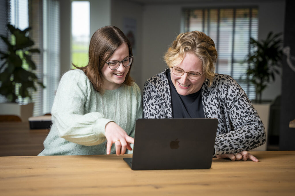 Twee vrouwen glimlachen en kijken naar een laptop op een houten tafel in een lichte kamer met planten en grote ramen.