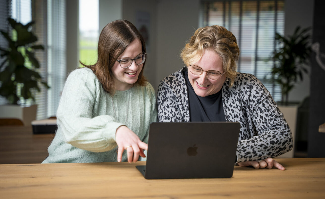 Twee vrouwen glimlachen en kijken naar een laptop op een houten tafel in een lichte kamer met planten en grote ramen.