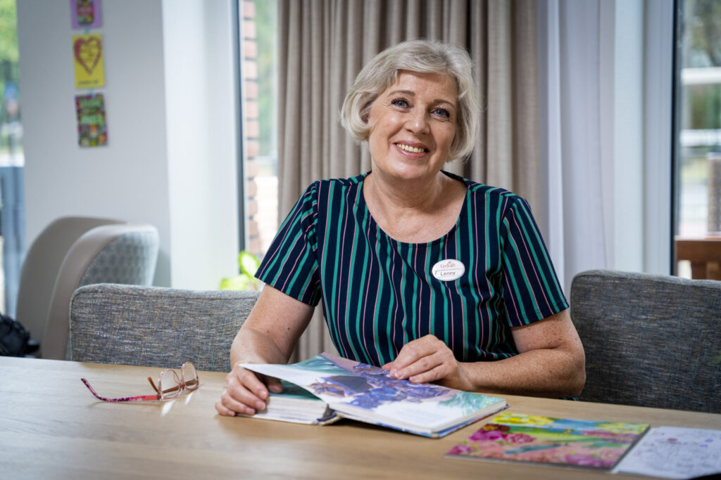 Vrouw met kort grijs haar zit aan tafel en bladert door een tijdschrift. Ze glimlacht en draagt een gestreepte jurk.