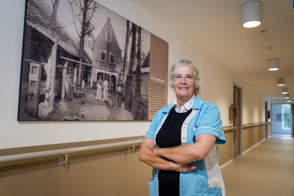 Vrouw in zorguniform staat met gekruiste armen in een gang, met een historische zwart-witfoto aan de muur achter haar.