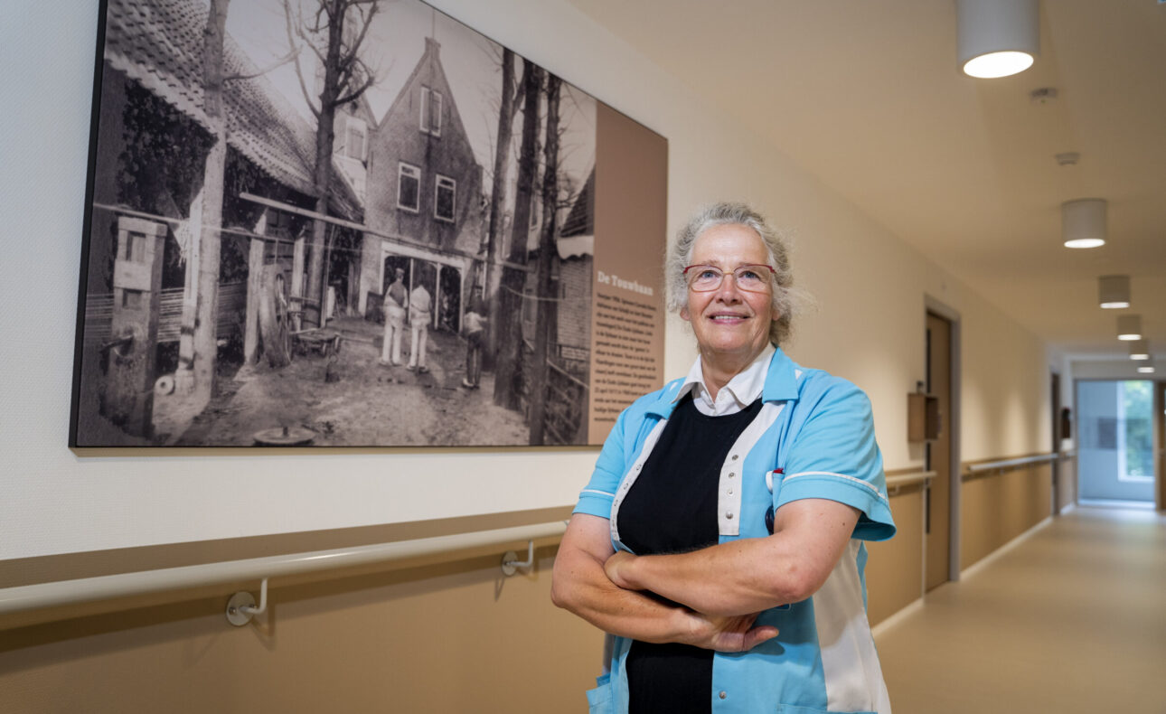 Vrouw in zorguniform staat met gekruiste armen in een gang, met een historische zwart-witfoto aan de muur achter haar.