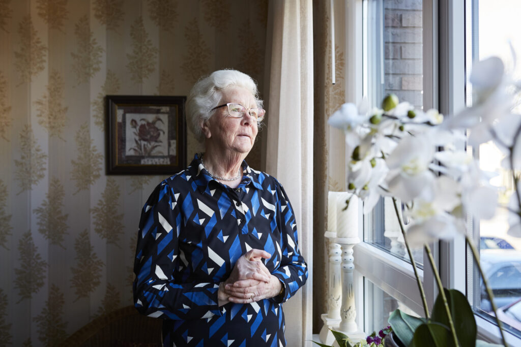 Een oudere vrouw met wit haar kijkt peinzend uit het raam. Ze draagt een blauw met zwart patroon blouse. Orchideeën op de voorgrond.