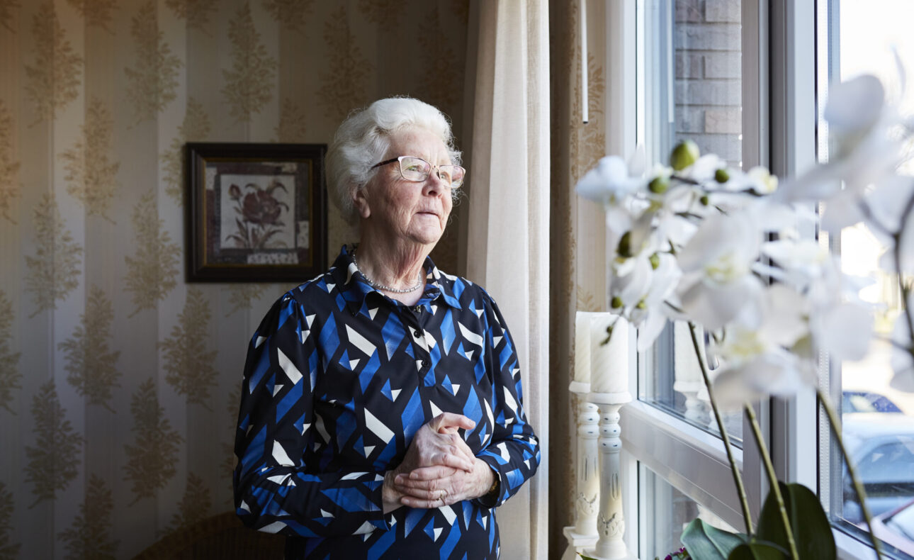 Een oudere vrouw met wit haar kijkt peinzend uit het raam. Ze draagt een blauw met zwart patroon blouse. Orchideeën op de voorgrond.