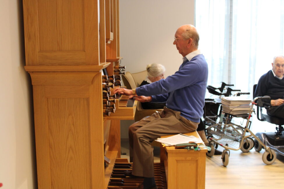 Een oudere man speelt orgel in een lichte kamer. Op de achtergrond zitten twee andere mensen, waarvan één in een rolstoel.