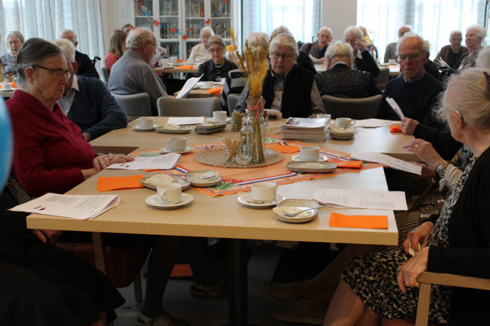 Een groep ouderen zit aan een tafel in een gemeenschappelijke ruimte, omringd door papieren en kopjes, in gesprek met elkaar.