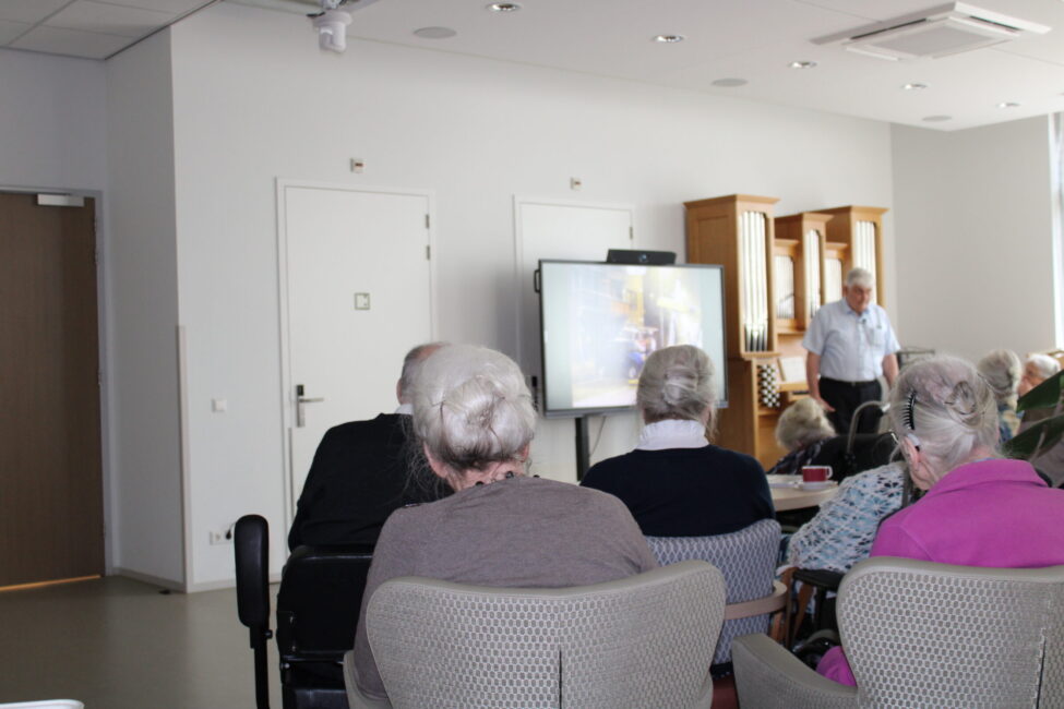 Een groep ouderen zit in een zaal en kijkt naar een presentatie op een scherm. Een man staat voor de groep en spreekt.