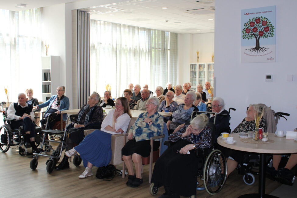 Groep ouderen zit in een lichte ruimte, aandachtig kijkend naar een presentatie. Sommigen zitten in rolstoelen.