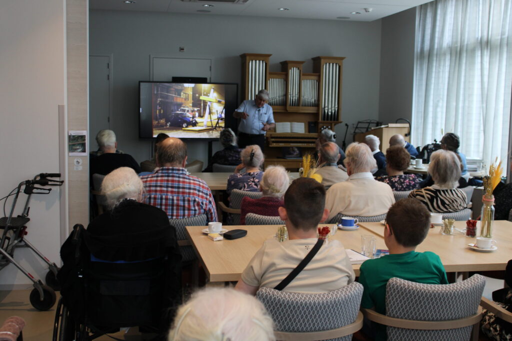 Een groep ouderen en enkele jongeren kijken naar een presentatie op een groot scherm in een gemeenschappelijke ruimte.