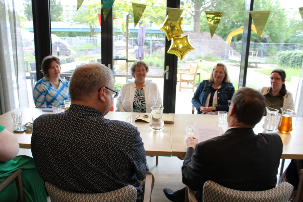 Een groep mensen zit aan een tafel in een feestelijk versierde ruimte met gekleurde vlaggen en gouden sterballonnen.