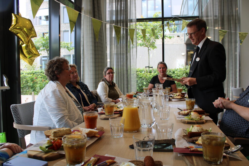 Een groep mensen zit aan een tafel met eten en drinken, terwijl een man in pak een toespraak houdt. Er hangen feestelijke vlaggen.