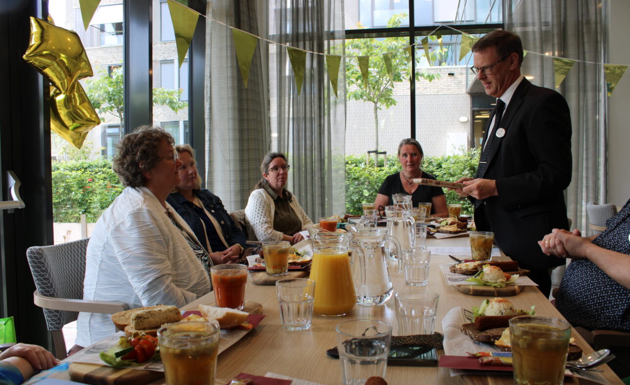 Een groep mensen zit aan een tafel met eten en drinken, terwijl een man in pak een toespraak houdt. Er hangen feestelijke vlaggen.