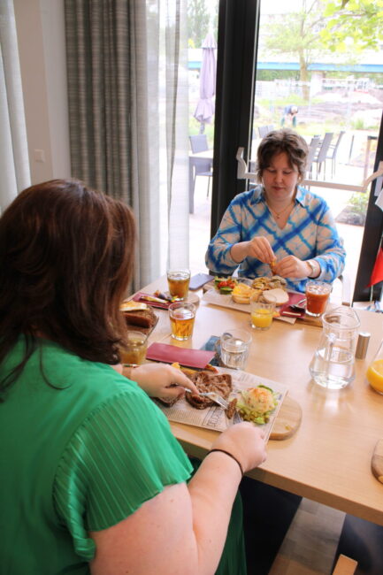 Twee vrouwen zitten aan een tafel in een restaurant en genieten van een maaltijd. Er staan drankjes en borden met eten op tafel.