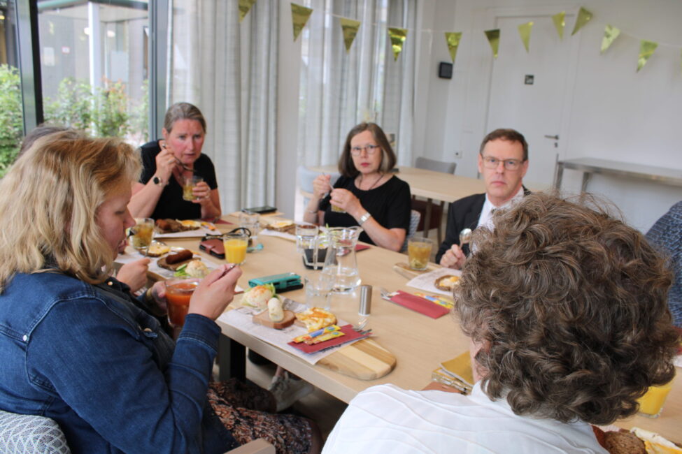 Groep mensen zit aan een tafel in een lichte ruimte, genietend van een maaltijd. Er hangen feestelijke vlaggetjes aan de muur.