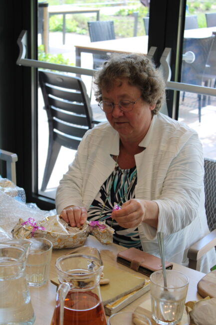 Een vrouw met krullend haar zit aan een tafel en werkt aan een handwerkproject. Er staan glazen en een kan op tafel.