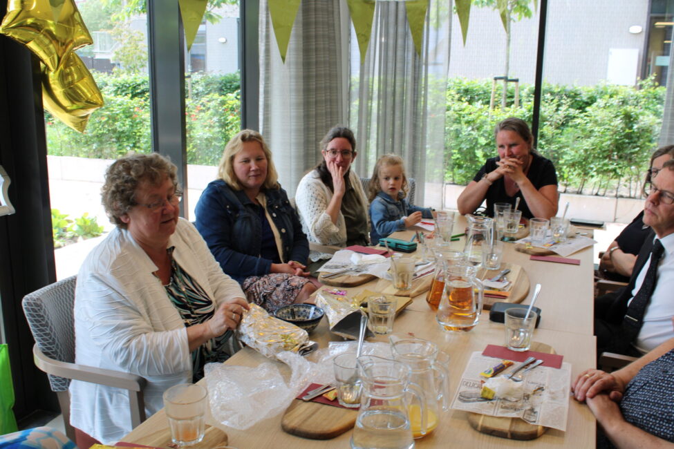 Groep mensen zit aan een tafel in een feestelijke ruimte, met ballonnen en decoraties, genietend van een maaltijd en elkaars gezelschap.