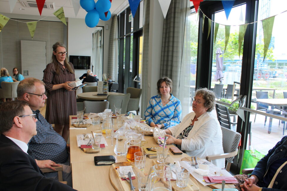 Een groep mensen zit aan een tafel in een feestelijk versierde ruimte met vlaggetjes en ballonnen, in gesprek met elkaar.