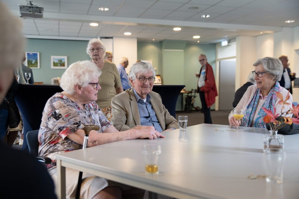 Een groep oudere mensen zit aan een tafel in een lichte ruimte tijdens een jubileumbijeenkomst, pratend en genietend van drankjes.