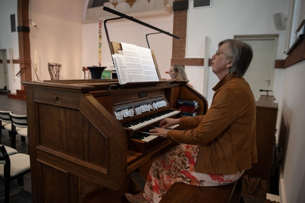 Een vrouw speelt orgel in een kerk tijdens een jubileumbijeenkomst. Ze draagt een bruine jas en een kleurrijke rok.