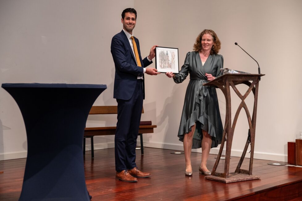 Twee mensen staan op een podium. De man in pak overhandigt een ingelijste foto aan de vrouw in een groene jurk.