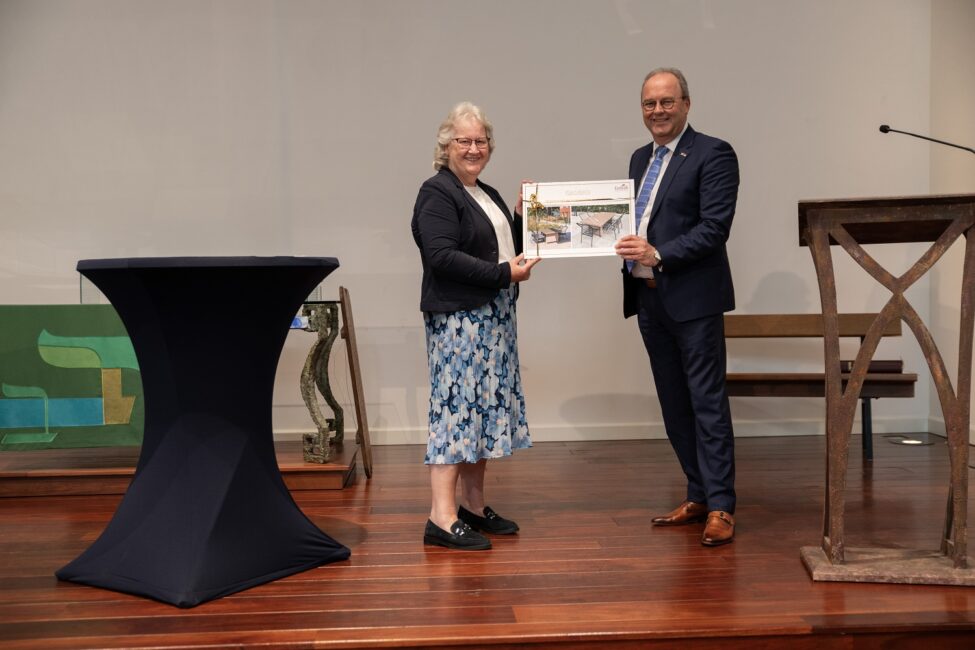 Twee mensen staan op een podium. Ze houden samen een ingelijste foto vast tijdens een jubileumbijeenkomst.