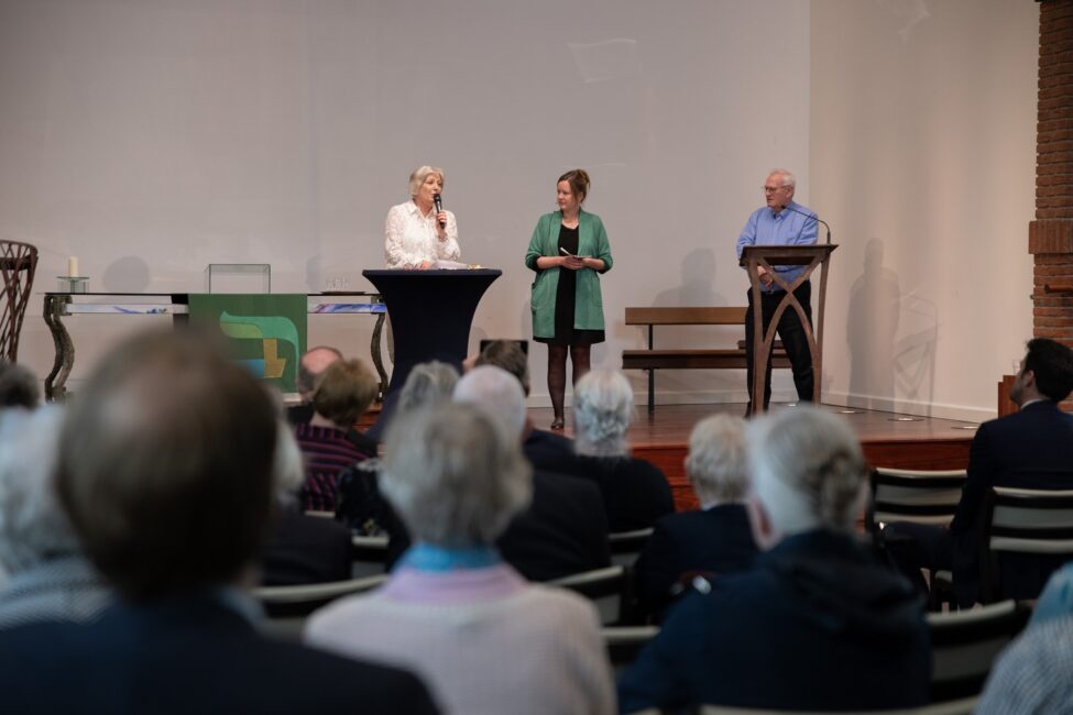 Een groep mensen zit in een zaal tijdens een jubileumbijeenkomst. Drie sprekers staan op het podium en praten met het publiek.
