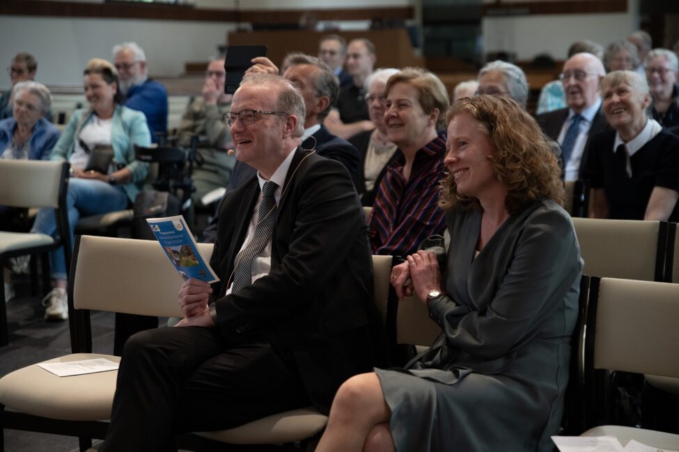 Een groep mensen zit in een zaal tijdens een jubileumbijeenkomst. Een man en een vrouw op de voorgrond lachen en kijken naar voren.