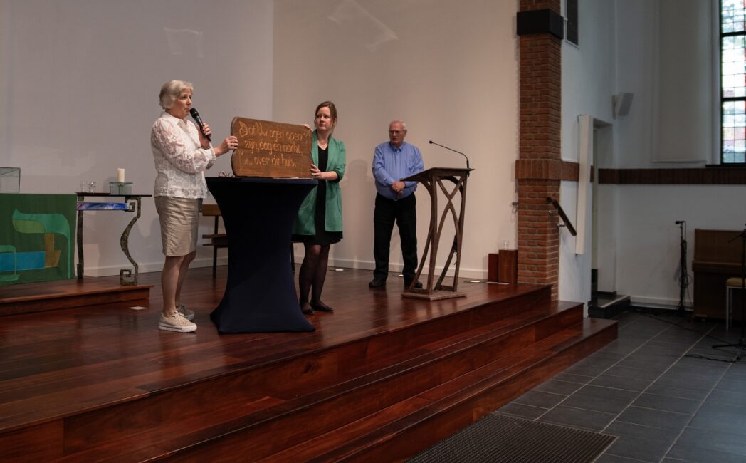 Drie mensen staan op een podium in een zaal. Een vrouw houdt een houten object vast terwijl een andere vrouw spreekt.