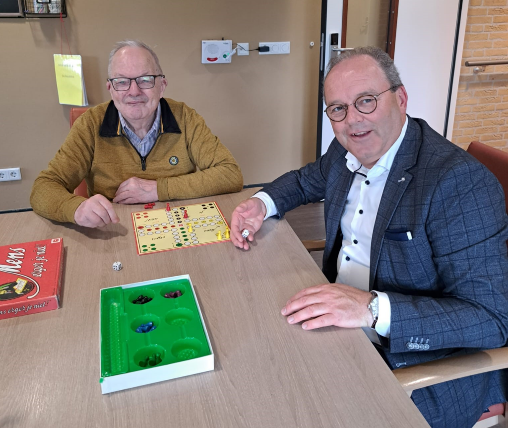 Twee mannen spelen een bordspel aan een tafel in een gezellige kamer. Een van hen houdt een dobbelsteen vast en glimlacht.