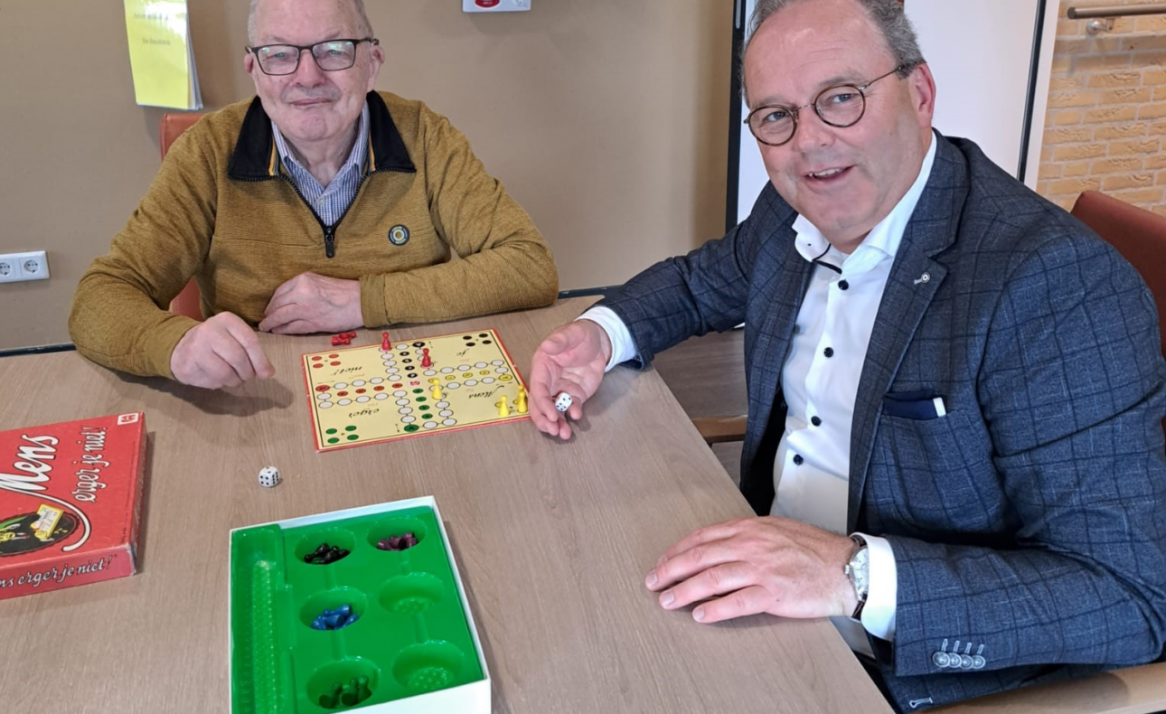 Twee mannen spelen een bordspel aan een tafel in een gezellige kamer. Een van hen houdt een dobbelsteen vast en glimlacht.