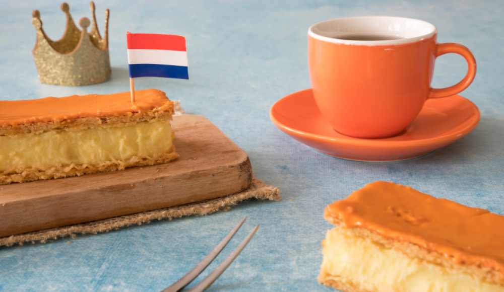 Oranje tompouce op een bord met een Nederlandse vlag, naast een oranje kopje koffie en een gouden kroon op een blauwe tafel.