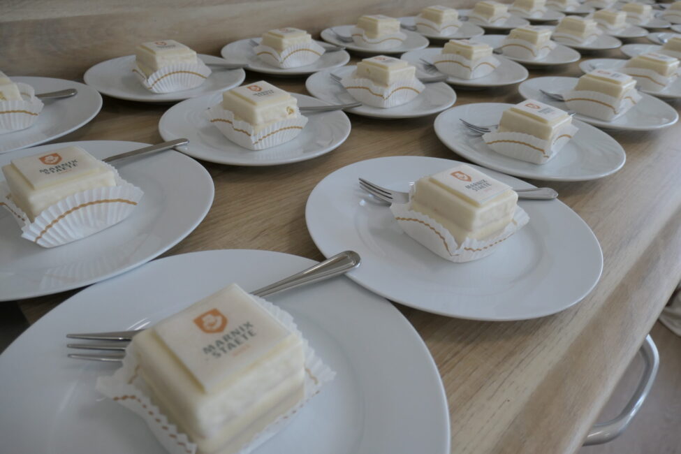 Rij witte gebakjes met logo op witte borden, elk met een vork, op een houten tafel. De gebakjes zijn vierkant en gelijkmatig verdeeld.