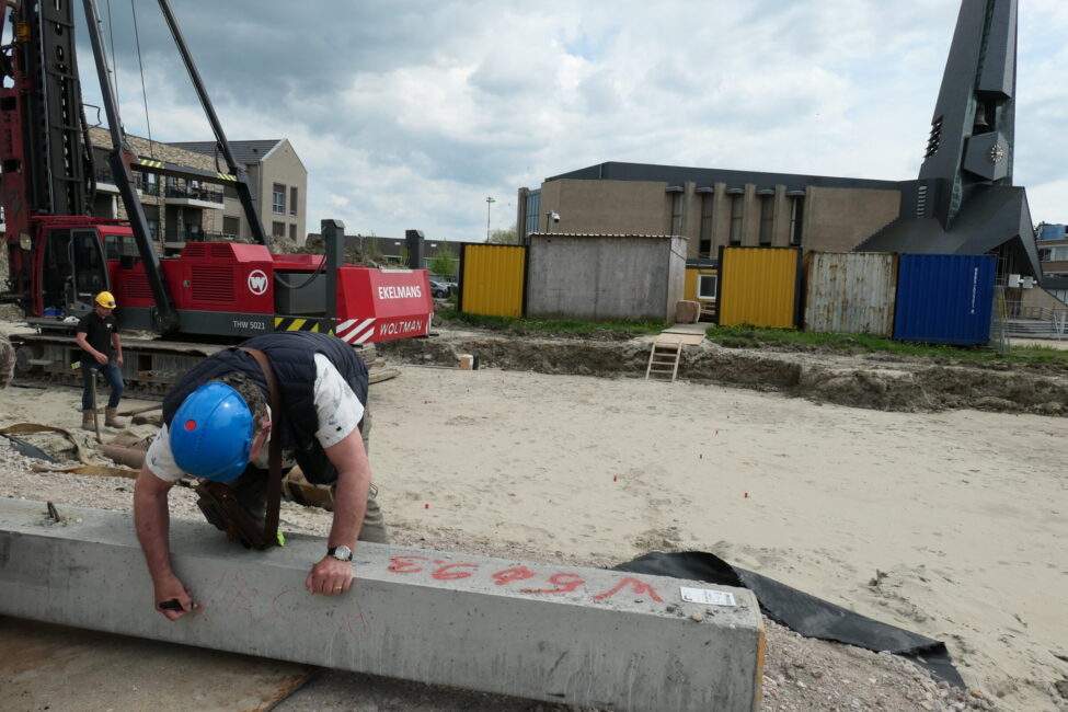Bouwvakker met blauwe helm werkt aan betonnen balk op bouwplaats, met hijskraan en gebouwen op de achtergrond onder bewolkte hemel.