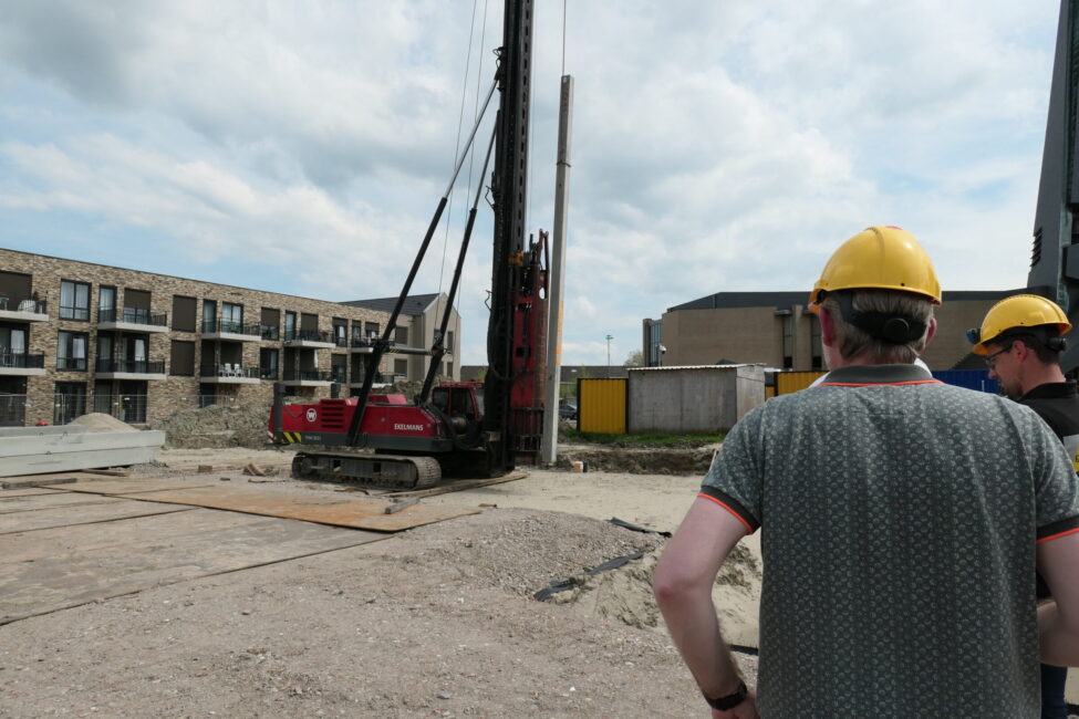 Twee mannen met gele helmen kijken naar een grote machine op een bouwplaats, omringd door moderne gebouwen en wolken.