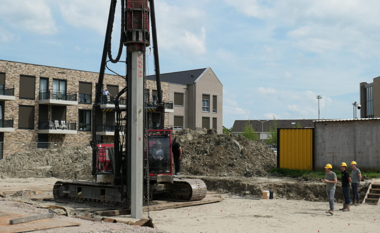 Bouwplaats met een heistelling en drie bouwvakkers in gesprek. Op de achtergrond zijn moderne appartementen te zien.