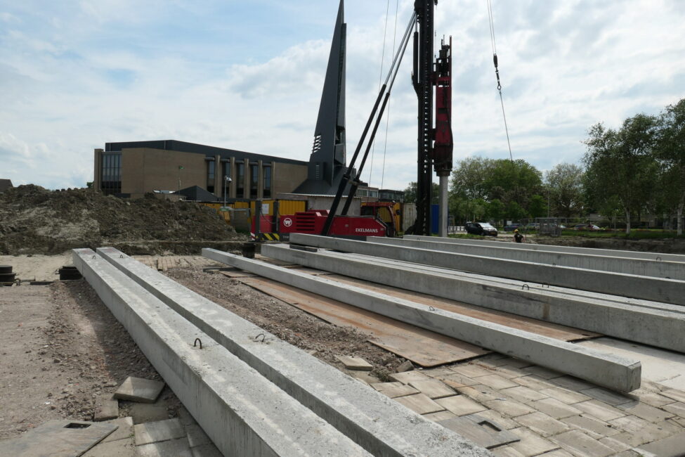 Bouwplaats met lange betonnen balken op de grond, een hijskraan en een kerk op de achtergrond onder een bewolkte hemel.