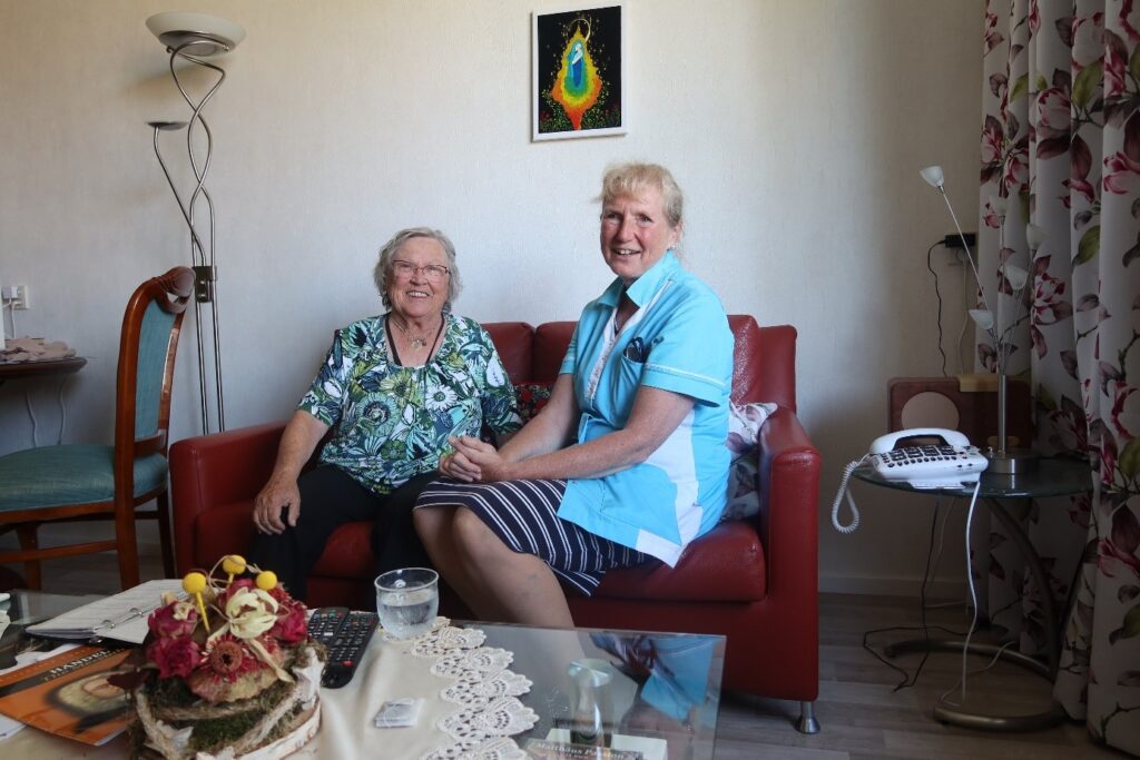 Twee vrouwen zitten glimlachend op een rode bank in een woonkamer. Er staat een telefoon op een tafel naast hen.