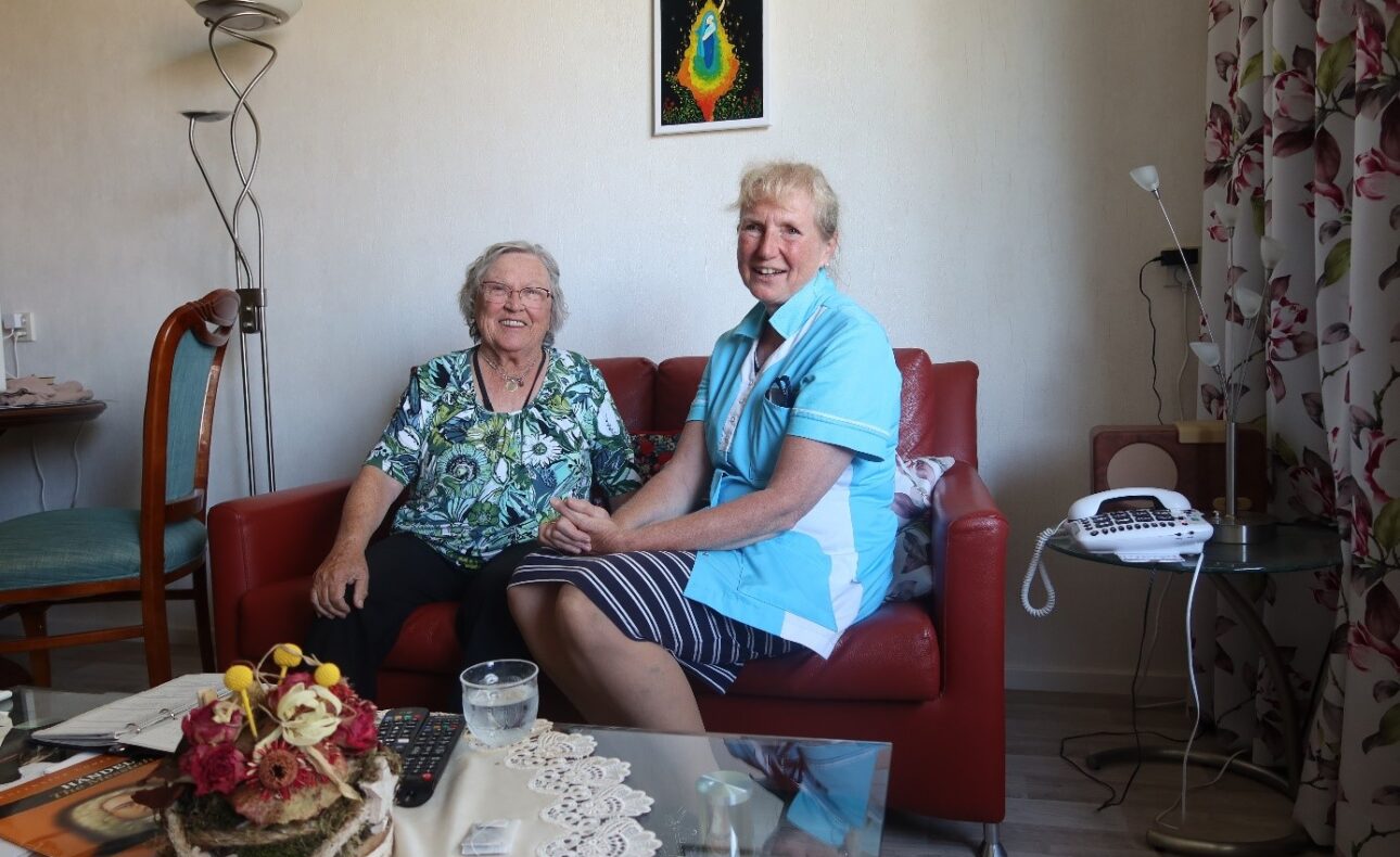 Twee vrouwen zitten glimlachend op een rode bank in een woonkamer. Er staat een telefoon op een tafel naast hen.