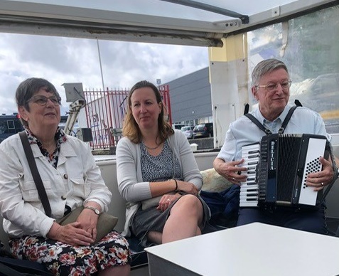 Drie mensen zitten samen op een boot. De man rechts speelt accordeon. De lucht is bewolkt en er zijn gebouwen op de achtergrond.