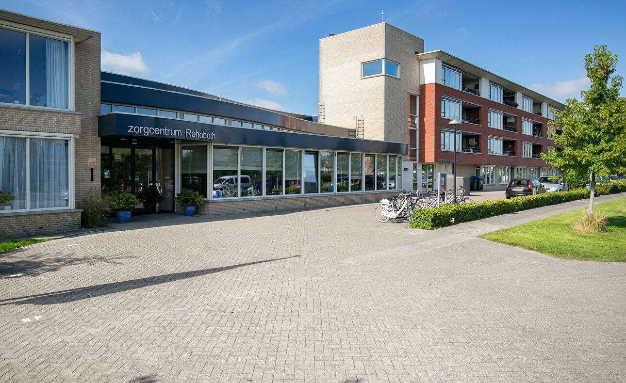 Afbeelding van de buitenkant van een modern zorgcentrum met grote ramen, een bakstenen gevel en een betegelde oprit.