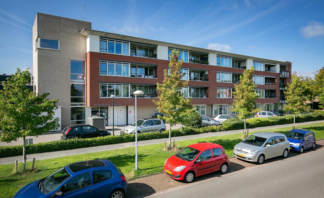 Appartementencomplex met vier verdiepingen, omgeven door bomen en geparkeerde auto's langs de straat op een zonnige dag.