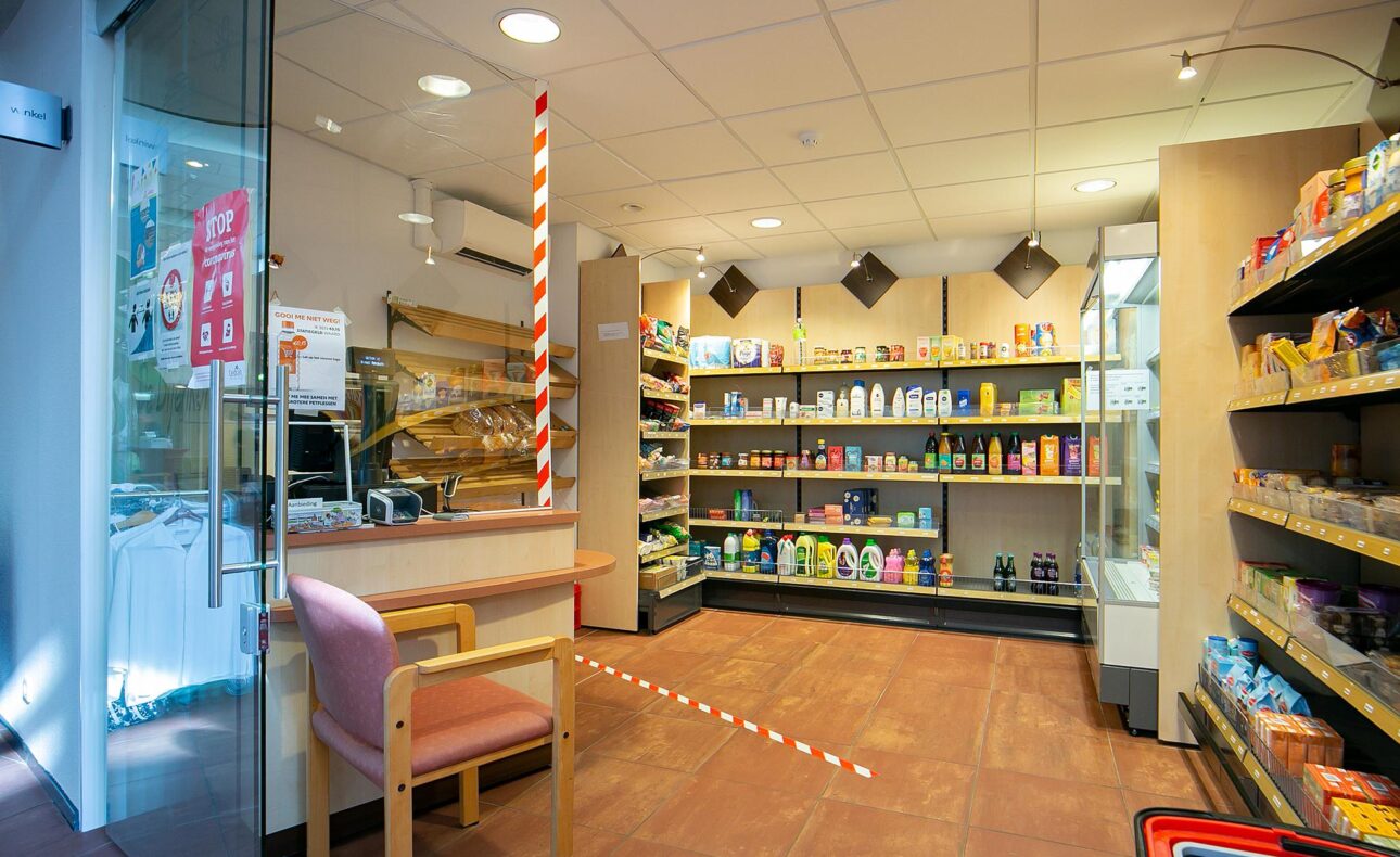 Een kleine winkel met houten schappen vol producten, een roze stoel bij de ingang en een glazen deur met posters.
