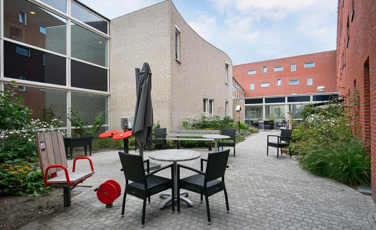 Binnentuin met tafels en stoelen, omgeven door moderne bakstenen gebouwen en groene planten onder een blauwe lucht.