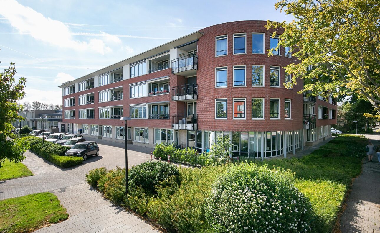 Afbeelding van een modern, rood bakstenen gebouw met meerdere verdiepingen, omgeven door groen en een zonnige blauwe lucht.
