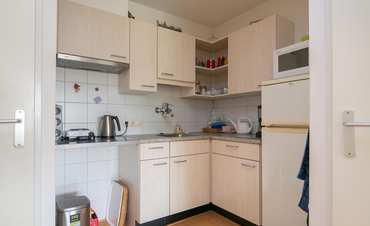Kleine keuken met houten kastjes, een magnetron, waterkoker en koelkast. Er zijn witte tegels op de muur en een gootsteen.