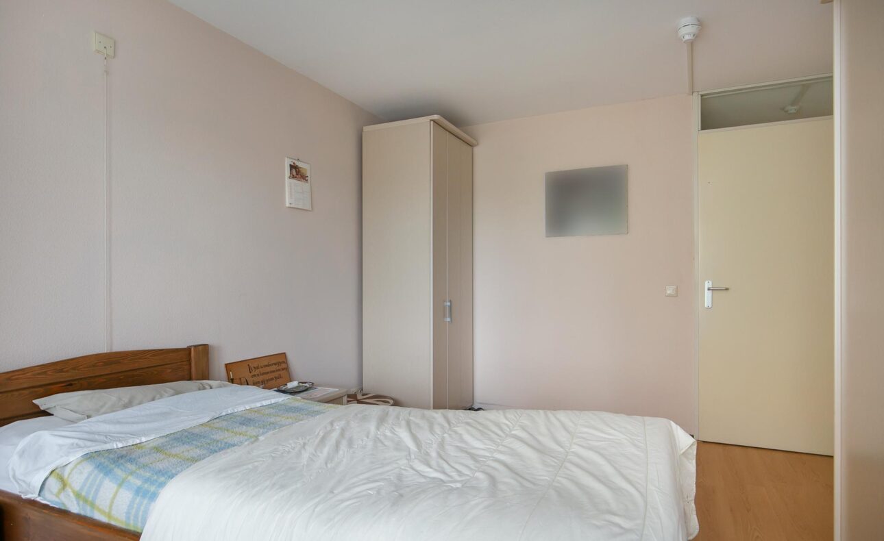Een eenvoudige slaapkamer met een houten bed, wit beddengoed, een kast en een stoel tegen een lichte muur.