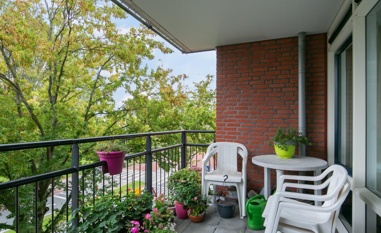 Balkon met groene planten en kleurrijke bloemen in potten, witte plastic stoelen en een tafel, met uitzicht op bomen.